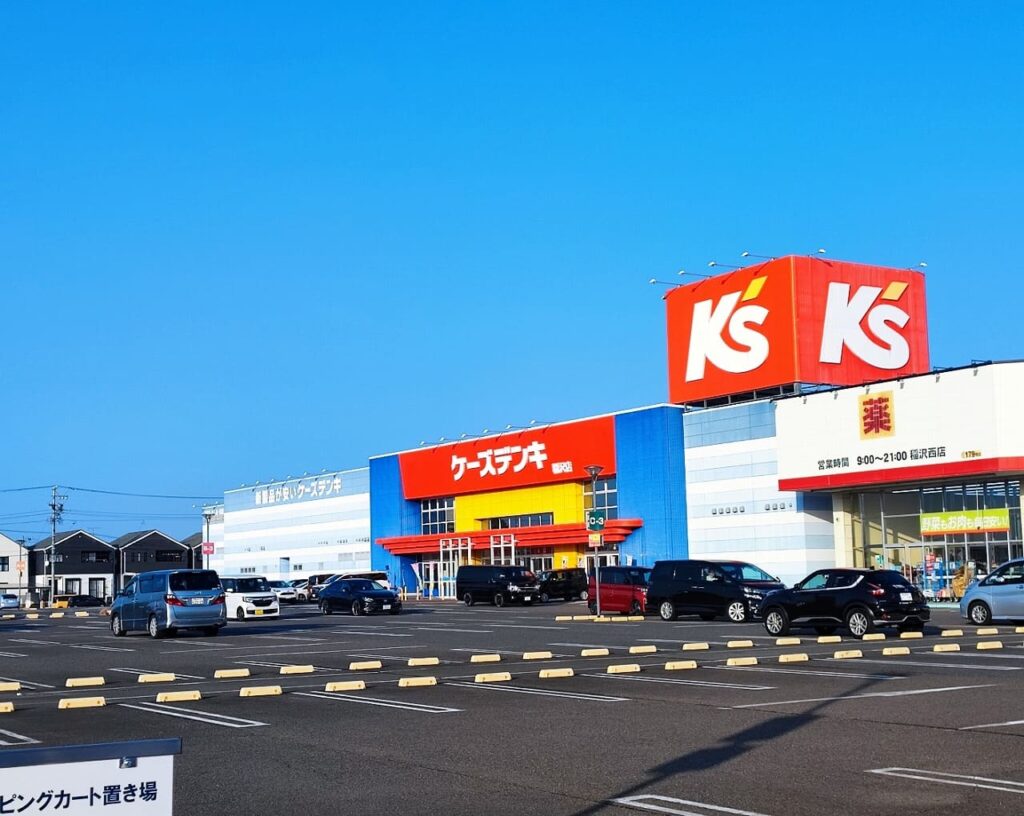 ニッケタウン稲沢のケーズデンキ稲沢店との外観。赤と青を基調とした大型家電量販店の建物で、広い駐車場には多数の車両が停車。晴天の青空の下で撮影された施設全景