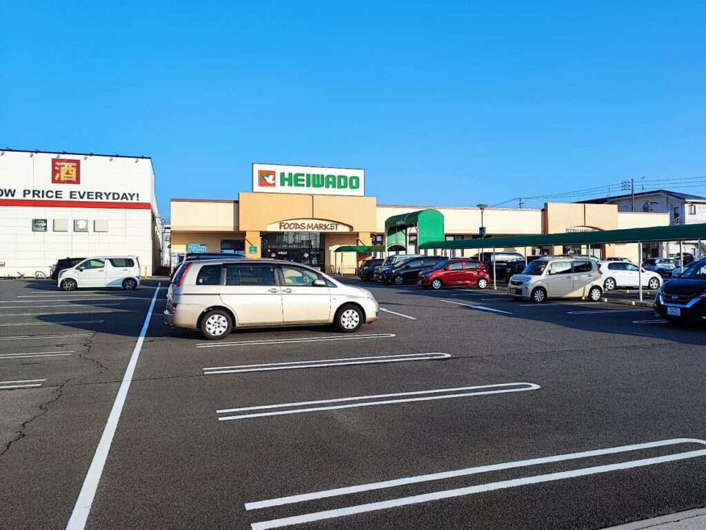 ニッケタウン稲沢の平和堂フーズマーケットの外観。緑色の平和堂ロゴが目立つクリーム色の建物で、左側には酒類販売の看板も見える。駐車場には複数の車両が停車し、日常的な買い物客で賑わう様子