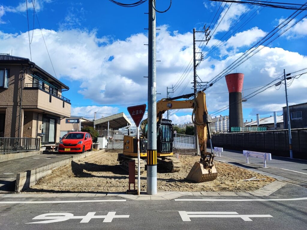 愛知県稲沢市奥田大沢町にある売地の現地写真。前面道路に接する整地済みの敷地で、重機が設置されている。交差点に面した角地で、周囲には住宅や工場が立ち並び、道路幅員と見通しの良さが確認できる。