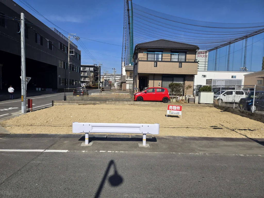 愛知県稲沢市奥田大沢町の売地現地写真。整地済みの約39坪の土地を前面道路から撮影しており、敷地内に売物件の看板が設置されている。周囲には住宅や事業所があり、平坦で建築しやすい更地の様子が分かる。