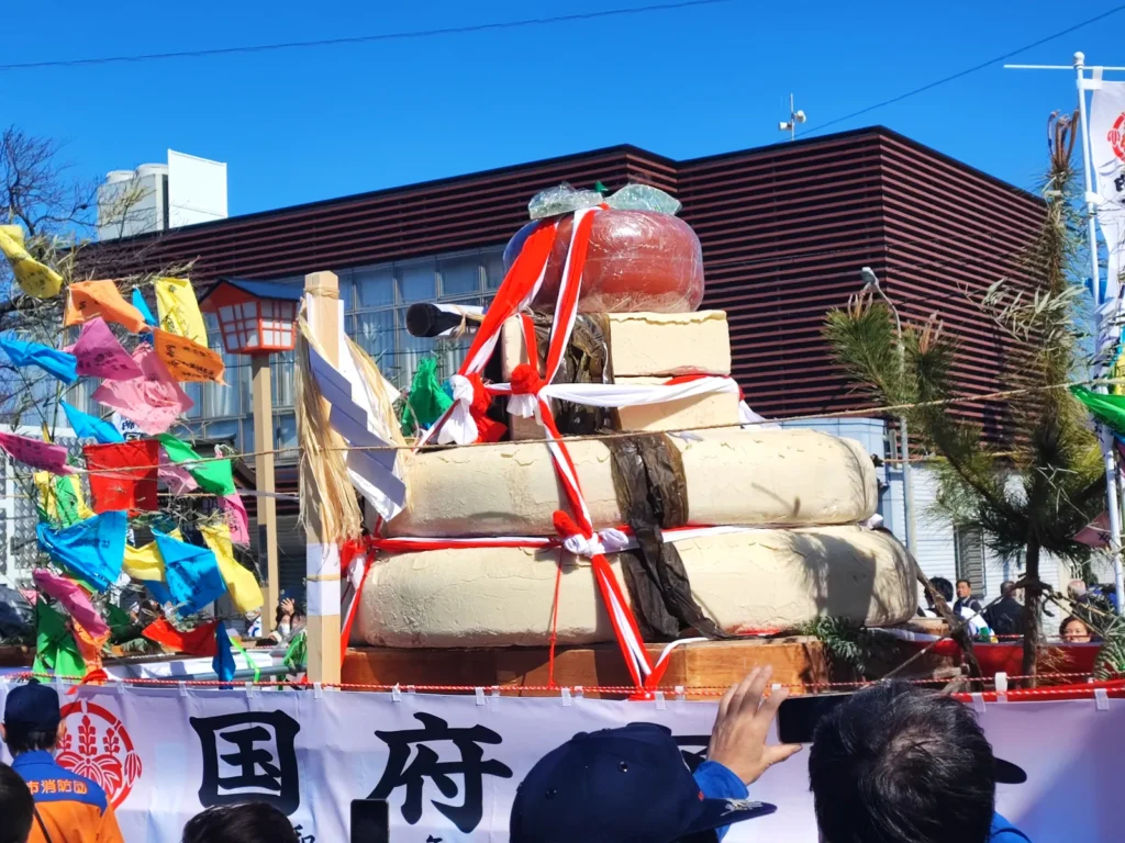 トラックの荷台に鎮座する、三段重ねの巨大な大鏡餅のクローズアップ。一番上には大きなダイダイが載り、餅の表面には乾燥によるひび割れが見える。周囲には魔除けの「なおいぎれ」が風に棚引いている。