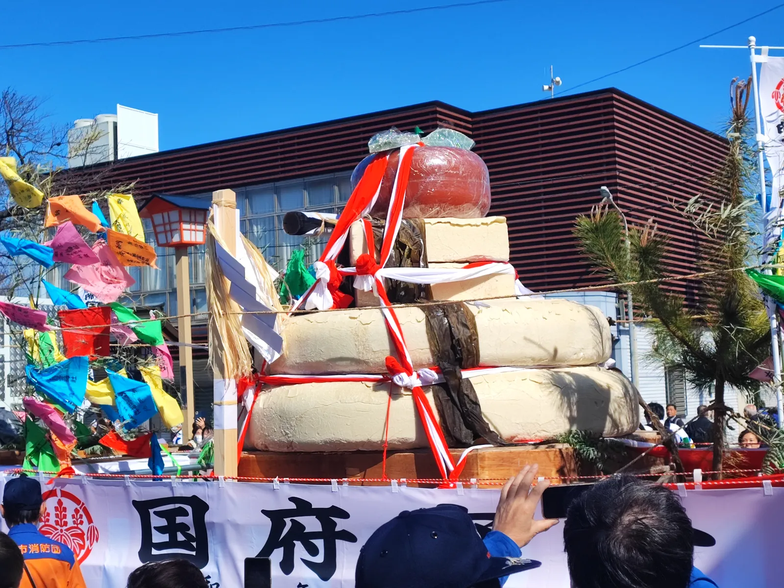 トラックの荷台に鎮座する、三段重ねの巨大な大鏡餅のクローズアップ。一番上には大きなダイダイが載り、餅の表面には乾燥によるひび割れが見える。周囲には魔除けの「なおいぎれ」が風に棚引いている。