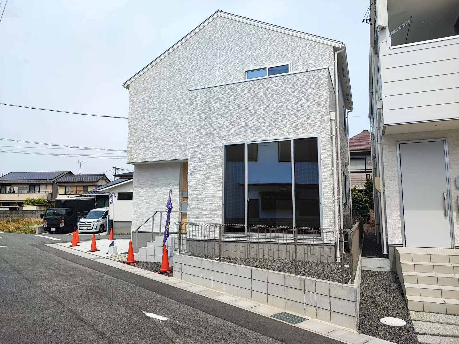 未入居の中古一戸建の外観写真。白を基調としたシンプルな2階建て住宅で、大きな窓から採光が取れる設計。前面道路に面し、敷地は整備されており駐車スペースあり。周辺は戸建住宅が並ぶ落ち着いた住宅街の環境。