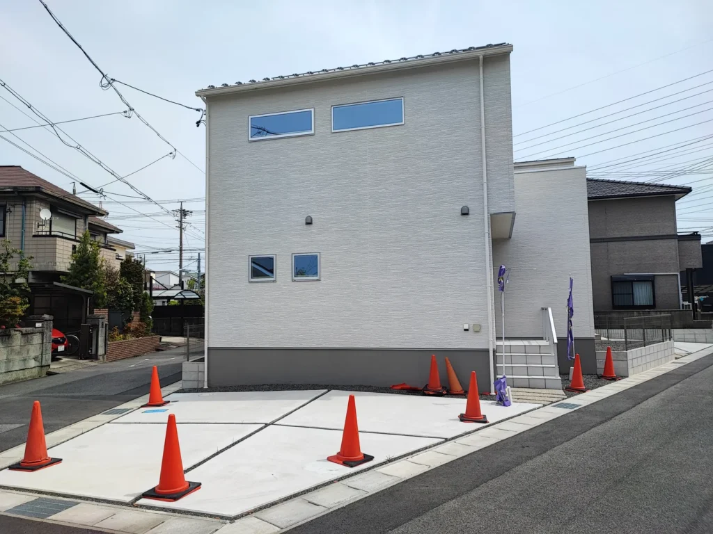 未入居の中古一戸建の外観写真。白い外壁の2階建て住宅で、建物正面に広いコンクリート舗装の駐車スペースがあり、2台以上駐車可能。前面は道路に面した角地状の開放感のある立地で、周囲は戸建住宅が並ぶ落ち着いた住宅街。