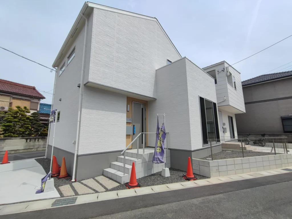 未入居の中古一戸建の外観と玄関まわりの写真。白い外壁の2階建て住宅で、玄関まで階段と手すりが設置されている。玄関横には大きな窓があり採光が確保され、前面には駐車スペースと道路に面した開放的な立地。周囲は戸建住宅が並ぶ静かな住宅街。