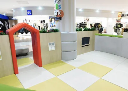 アピタタウン稲沢の館内に設置された無料の屋内キッズスペース「キッズパーク」。オレンジ色のアーチ型トンネルと黄色・白のタイル床、丸いクッション性の柱カバーが並び、小さな子どもが安全に遊べるよう配慮された造り。奥にはGUなど衣料品売場が見える。