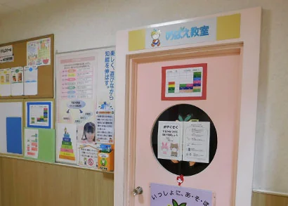 アピタタウン稲沢2階にある「めばえ教室」の入口。ピンク色の扉に「めばえ教室」のロゴプレートと、親子で参加できる催しの案内チラシが掲示されている。壁には各種イベント告知やお知らせの掲示板が並ぶ。