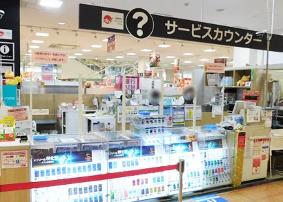 アピタタウン稲沢1階のサービスカウンター前の様子。案内表示の下にATMコーナーが並び、両替機・ATM利用の案内ポスターが掲示されている。買い物客のプライバシーに配慮し人物部分はぼかされている。手前には携帯電話ケースなどの商品棚が写る。