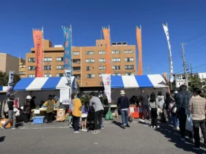 びさいまつりの会場周辺の様子。青と白のテントが並ぶ露店の背後に「びさいまつり」と書かれたオレンジと白ののぼり旗が複数掲げられ、奥には尾西庁舎の茶色いビルが見える。手前には来場者が行き交う晴天の屋外風景。