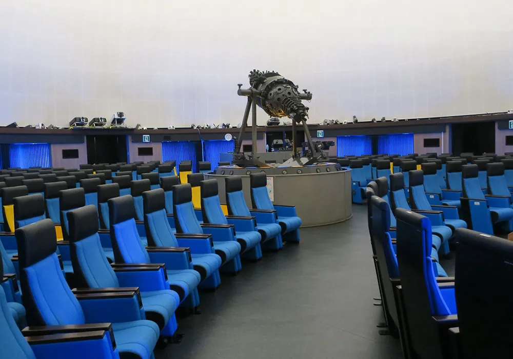 一宮地域文化広場プラネタリウム館のドーム内観。青い座席が扇状に何列も並び、中央通路の先に恒星投影機が据えられている。壁際には青い間接照明が連なり、天井は白いドームスクリーンになっている。投映前で座席はすべて空席。
