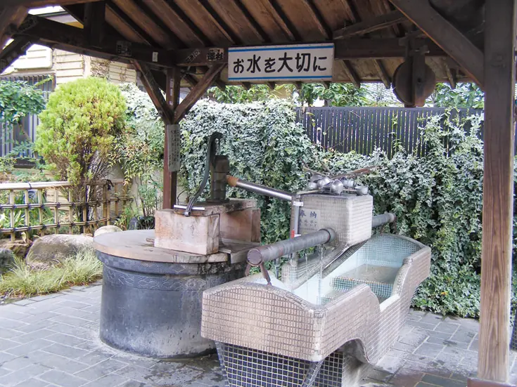 矢合観音の境内にある井戸の手押しポンプと水汲み場。木造の屋根の下に古い石造りの井戸とポンプ、水を受ける石造りの水盤が設置され、「お水を大切に」と書かれた案内板が掛かっている。背景には緑の生垣が茂っている。
