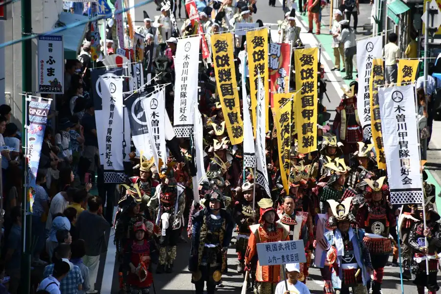 商店街を戦国武将姿の行列が練り歩く一豊まつりの戦国時代パレード。『戦国武将団 山内一豊家臣団』と書かれた立て札を先頭に、甲冑姿の武将たちが列をなし、沿道には見物客が並び、黄色や白の幟旗が何本も掲げられている。