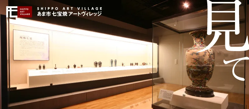 あま市七宝焼アートヴィレッジの常設展示室。壁沿いのガラスケースには「尾張七宝」の解説パネルと、七宝焼の花瓶や小皿が横一列に並ぶ。手前には花鳥模様が描かれた大きな花瓶が単独のガラスケースに納められ、スポットライトで照らされている。木目調の床の落ち着いた展示室。