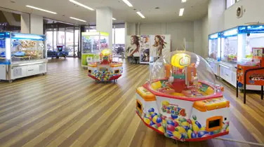 稲沢グランドボウル館内のゲームコーナー。クレーンゲームやカプセルトイの機械が木目調の床に並び、ガラス越しに明るい入口側の光が差し込んでいる。ボウリングの合間に子どもが遊べるスペース。