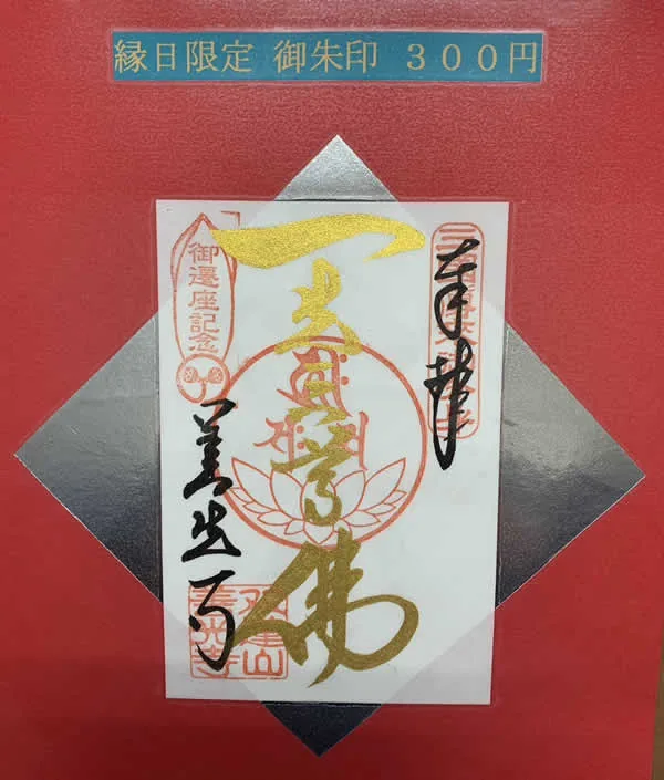 赤色の台紙に「縁日限定 御朱印 300円」と書かれた案内の下に、実際にいただける御朱印の写真。白い和紙に金色の文字で「南無阿弥陀仏」と大きく書かれ、蓮の花をかたどった朱印と複数の朱印が押されている。人物は写っていない。