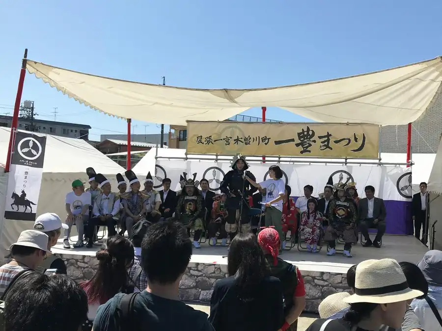 『尾張一宮木曽川町 一豊まつり』と大きく書かれた黄色い横断幕の下、甲冑姿の武将役や着物姿の千代役が特設ステージに並んで座り、白い日よけテントの下で来場者が後ろ姿でステージを見守っている一豊まつりの会場の様子。