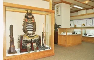 清洲城文化財収蔵庫の展示室。ガラスケースの中に、兜と胴を備えた甲冑一式と長い槍・刀が展示されている。木目調の展示台と照明の下、奥にも別の展示ケースが並び、戦国時代の武具を間近に見られる室内の様子。