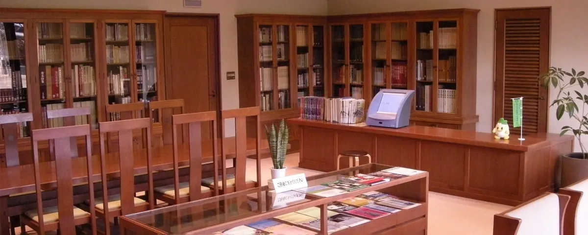 荻須記念美術館内の閲覧コーナー。木製の書棚に美術関連の書籍がびっしりと並び、木製の椅子とテーブル、ガラスケースに入った図録やパンフレットが置かれている。壁際には検索用の端末と観葉植物も見える。