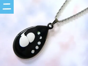 しずく形の黒い台に、白い雲のような模様と小さな水玉が並ぶ七宝焼のペンダントトップ。銀色のチェーンが付き、淡いピンクとブルーの背景の上に置かれている。体験教室で作れる作品の一例。