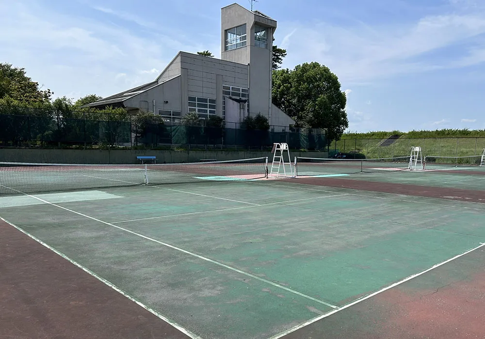 尾西文化広場のテニスコートを写した写真。奥にふれあい会館の展望塔が見え、手前に緑と赤茶色に塗り分けられたハードコート3面、白いラインとネット、審判台が並んでいる。晴天で青空と白い雲が広がり、コートの周囲は緑のフェンスで囲まれている。