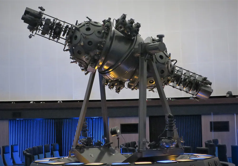 一宮地域文化広場プラネタリウム館の投影機「GMⅡ-18-AT」のクローズアップ。左右に恒星球を持つツインボール型の光学式プラネタリウム投影機で、球面には多数の小型レンズが放射状に取り付けられている。背景に白いドームスクリーン、下方には青い座席がうっすら見える。