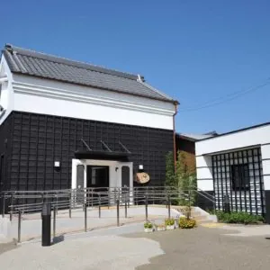 津島市観光交流センターの敷地内にある土蔵造りの建物。黒い板張りの外壁と白い漆喰の窓枠を持つ伝統的な土蔵で、瓦屋根が乗り、入口には木製の看板と手すり付きのスロープが設置されている。手前には低い植え込みがある。