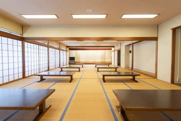 祖父江ふれあいの郷の和室休憩室の写真。畳敷きの広い部屋に、木製の座卓が両側に3台ずつ並んでいる。左側には障子窓が連なり、正面奥には一段高いスペースがある。天井には長方形の照明が3列並ぶ。人物は写っていない。
