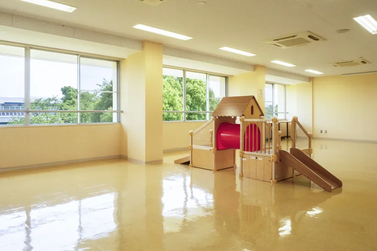 祖父江ふれあいの郷のプレイルームの写真。クリーム色の壁と大きな窓のある明るい部屋に、赤い屋根と滑り台のついた木製の子ども向け遊具が置かれている。床は光沢のあるベージュのタイルで、窓の外には緑の木々が見える。人物は写っていない。
