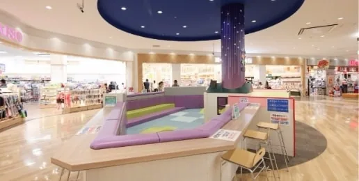 テラスウォーク一宮の館内にある円形のキッズスペース。紫と黄緑のクッション性ベンチが円を描くように配置され、中央には青い装飾柱が立つ。手前には木製のスツールが並び、奥には専門店の通路が続いている。