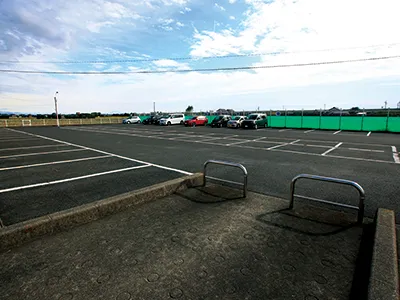 羽島温泉の駐車場。舗装されたスペースに白線の区画が引かれ、奥に数台の車が停まっている。手前には車止めの金属柵が2つ設置され、緑色のフェンス越しに周辺の建物が見える。