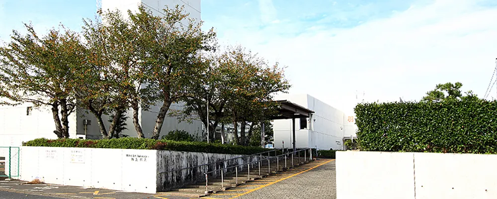 岐阜県羽島市桑原町にある羽島温泉の建物外観。白い壁の平屋の温泉施設の前に大きな木々が並び、駐車場から施設入口へ続く階段とスロープの手すりが見える。青空を背景に施設名の看板の一部が写り込んでいる。