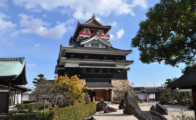 青空を背景にした清洲城天守閣の外観写真。黒漆塗りの下見板と白漆喰の壁を持つ4層の天守が石垣の上に建ち、最上階には赤い高欄が巡らされている。手前には庭石と植栽、右手に緑の大木が写る。