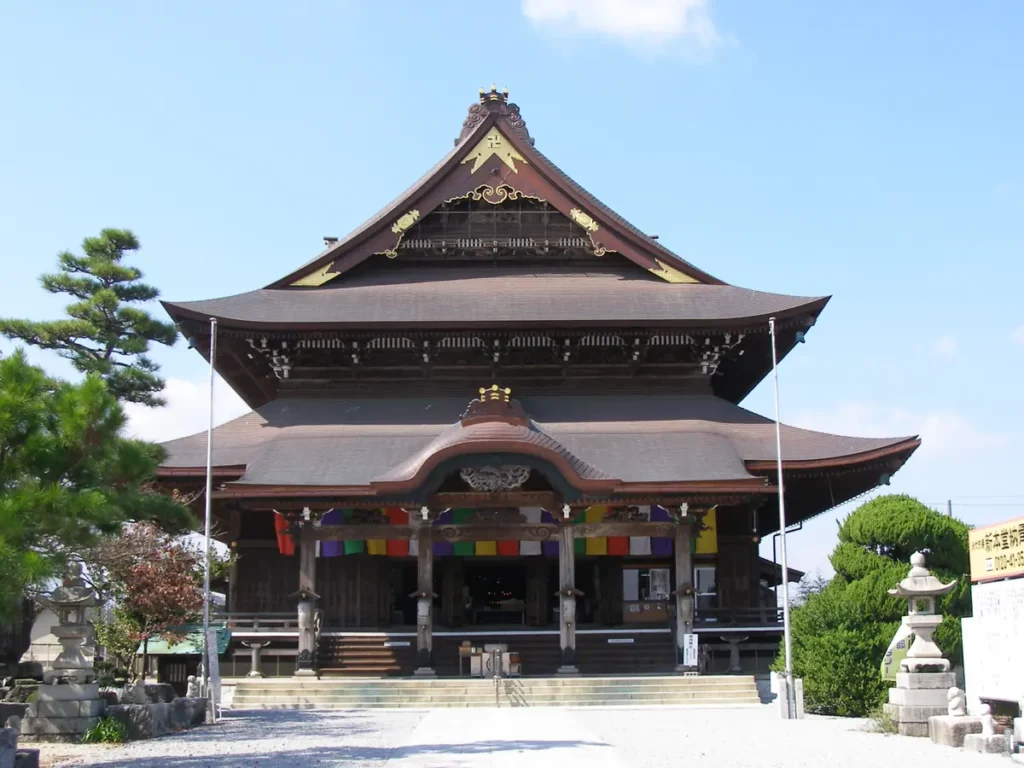 青空を背景に建つ善光寺東海別院の本堂正面。入母屋造りの大きな二重屋根と唐破風の向拝が特徴で、屋根には金色の飾り金具が施されている。手前に石灯籠と松の木、朱・黄・緑・白・紫の五色幕が本堂の柱に掛かる。参道は白い玉砂利敷き。人物は写っていない。