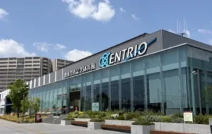エントリオ（豊田合成記念体育館）の外観。晴天の下、青い「ENTRIO」ロゴが入ったガラス張りの正面玄関。手前に植栽と歩道、右奥に高層マンションが見える稲沢駅前の街並み。