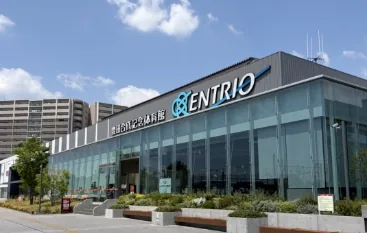 エントリオ（豊田合成記念体育館）の外観。晴天の下、青い「ENTRIO」ロゴが入ったガラス張りの正面玄関。手前に植栽と歩道、右奥に高層マンションが見える稲沢駅前の街並み。