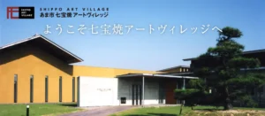 あま市七宝焼アートヴィレッジの建物外観。手前に大きな松の木、右手に灰色のコンクリート壁と木製の入口、左手にオレンジ色の外壁の建物が並ぶ。青空を背景にした施設のエントランス部分。