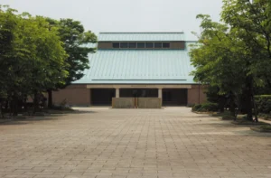 青緑色の大屋根が特徴的な荻須記念美術館の外観。左右対称の三角屋根とレンガ色の壁面、正面に石造りのモニュメントが据えられ、木々に囲まれた前庭の石畳が広がる稲沢市稲沢町の美術館正面写真。