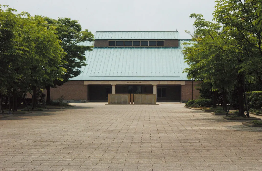 青緑色の大屋根が特徴的な荻須記念美術館の外観。左右対称の三角屋根とレンガ色の壁面、正面に石造りのモニュメントが据えられ、木々に囲まれた前庭の石畳が広がる稲沢市稲沢町の美術館正面写真。