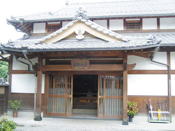 矢合観音の正面玄関。大きな瓦屋根と唐破風の屋根飾りが付いた木造建築で、格子戸の引き戸が開いており、軒下に「矢合観音」の文字が入った木製の看板が掲げられている。玄関脇には鉢植えの木や花、来訪者用の傘立てが置かれている。