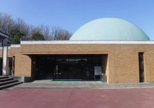 青みがかった半球形のドーム屋根が特徴的な一宮地域文化広場プラネタリウム館の外観。レンガタイル張りの平屋建物で、正面入口には「プラネタリウム館」の案内板があり、手前に赤茶色のタイル敷き広場と数段の階段、周囲には落葉した木々が広がる晴天の日中の写真。