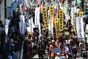 商店街を戦国武将姿の行列が練り歩く一豊まつりの戦国時代パレード。『戦国武将団 山内一豊家臣団』と書かれた立て札を先頭に、甲冑姿の武将たちが列をなし、沿道には見物客が並び、黄色や白の幟旗が何本も掲げられている。