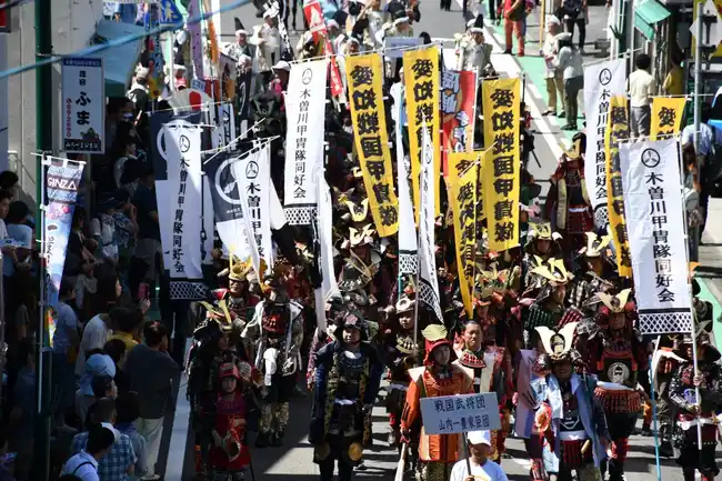 商店街を戦国武将姿の行列が練り歩く一豊まつりの戦国時代パレード。『戦国武将団 山内一豊家臣団』と書かれた立て札を先頭に、甲冑姿の武将たちが列をなし、沿道には見物客が並び、黄色や白の幟旗が何本も掲げられている。