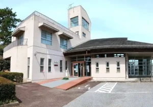 尾西文化広場のふれあい会館を正面から見た外観写真。淡いベージュ色の建物に四角い展望塔がそびえ、1階には自動ドアの入口とスロープ、車いす用駐車スペースの表示がある。左手前には松の木が植えられ、青空が広がっている。
