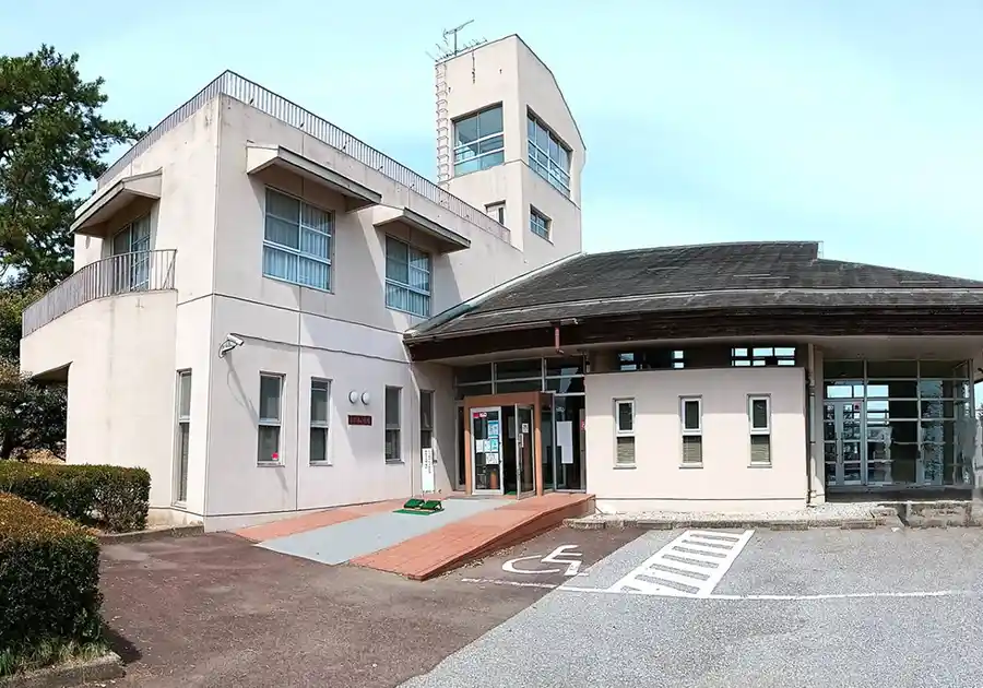 尾西文化広場のふれあい会館を正面から見た外観写真。淡いベージュ色の建物に四角い展望塔がそびえ、1階には自動ドアの入口とスロープ、車いす用駐車スペースの表示がある。左手前には松の木が植えられ、青空が広がっている。