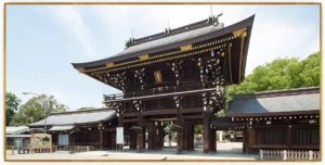 愛知県一宮市の真清田神社、正面から見上げた重厚な木造の楼門。軒先に白い紙垂（しで）状の装飾が連なり、左右に回廊と社殿が続く。晴れた青空と右手の緑の木々を背景にした写真。