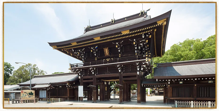 愛知県一宮市の真清田神社、正面から見上げた重厚な木造の楼門。軒先に白い紙垂(しで)状の装飾が連なり、左右に回廊と社殿が続く。晴れた青空と右手の緑の木々を背景にした写真。