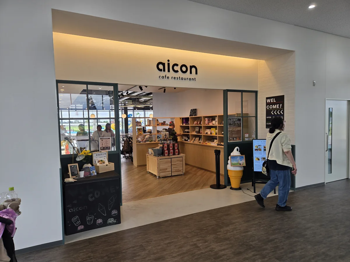 HASUパークのはす見テラス内にあるカフェレストランaiconの入口。木のルーバーと白い看板の奥に客席があり、平日の午後にも複数の来店客が食事や休憩をしている店内の様子。店内の明るさと利用時の雰囲気が伝わる一枚