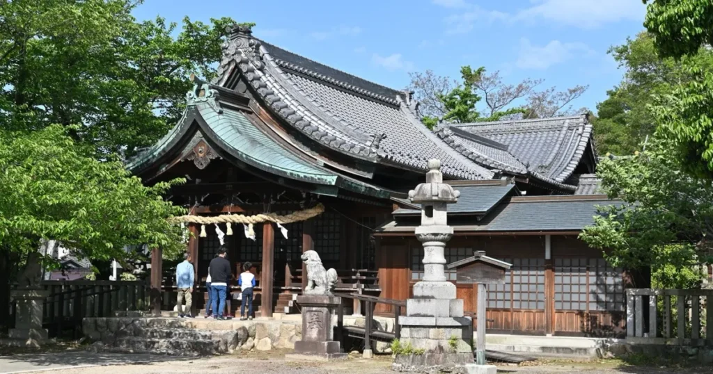 愛知県一宮市の石刀神社拝殿。緑青の唐破風屋根と重厚な瓦屋根を持つ木造社殿の前に、参拝者と石灯籠が写る写真。