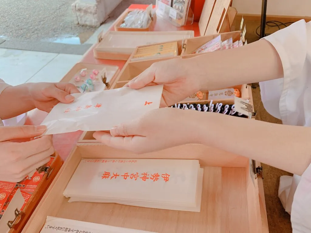 津島神社の授与所で、白い装束の巫女が参拝者に「悪疫退散門札」と朱字で書かれた白い和紙のお札を両手で手渡している様子。手元を大きくクローズアップした構図で、背景には木箱に並んだ神札やお守り、朱色の授与品コーナーが写り込んでいる。
