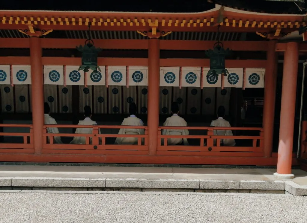 津島神社の祭典で、白い装束と黒い烏帽子を身につけた神職5人が、朱塗りの社殿の縁側に横一列に並んで座り、参道の方を向いて祭典に臨んでいる後ろ姿。軒先には青銅色の吊り灯籠が2つ下がり、白地に神紋を染め抜いた幕が柱の間に掛けられている。